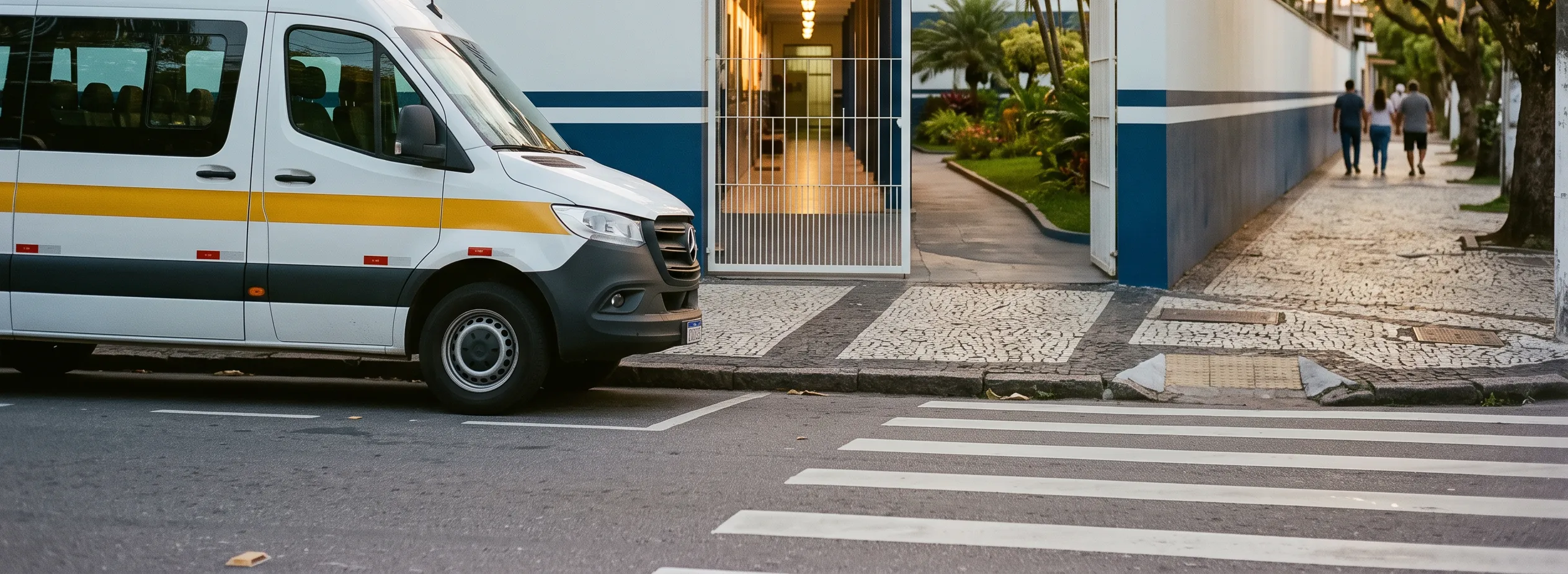 Uma van escolar branca com faixa amarela parada junto ao meio-fio, diante do portão aberto de uma escola, com o corredor interno iluminado ao fundo e uma faixa de pedestres em primeiro plano.