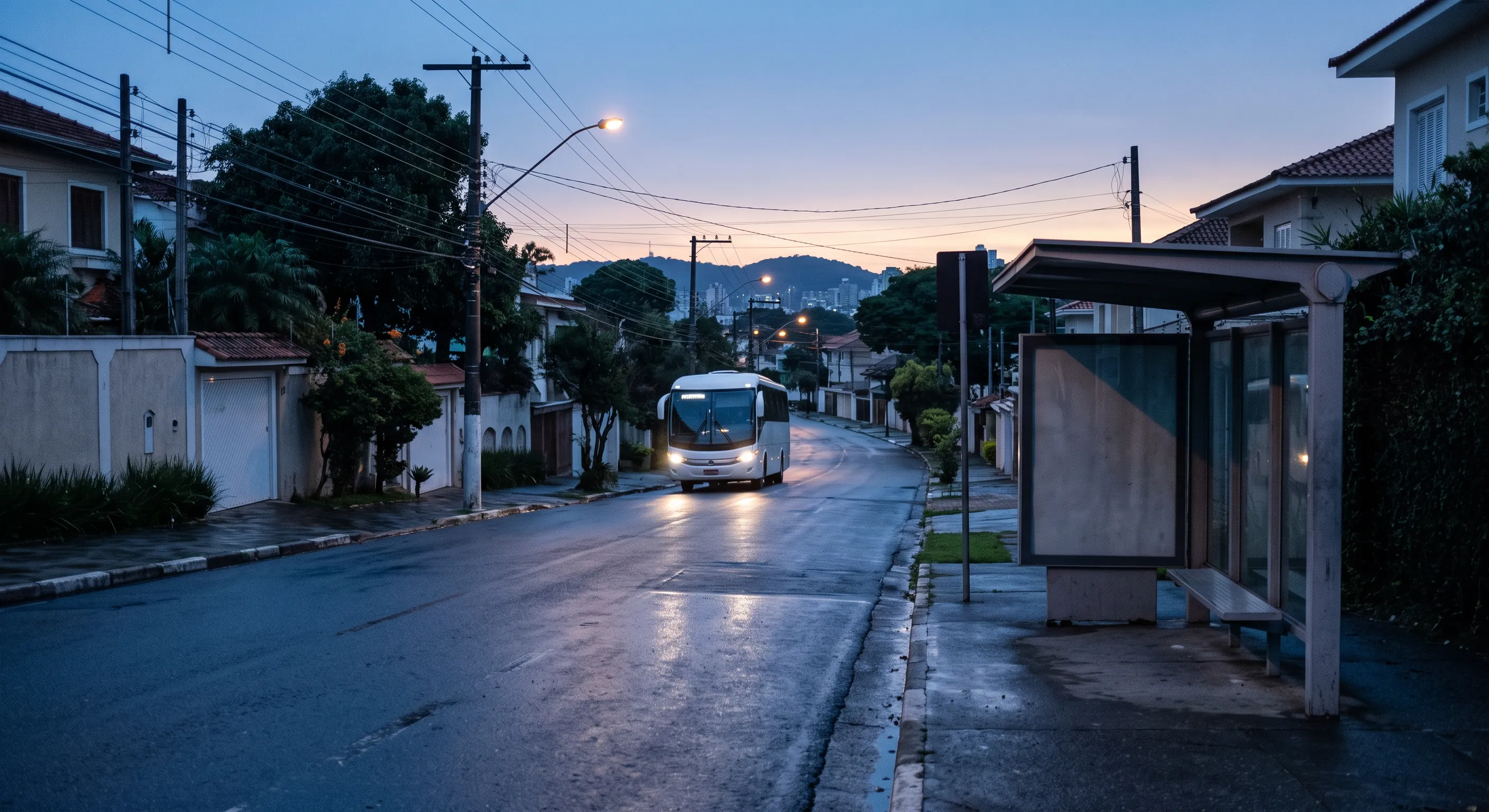 Rua residencial ao amanhecer, com o abrigo de um ponto de ônibus vazio em primeiro plano e um ônibus rodoviário de faróis acesos chegando ao fundo.